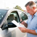 Un homme examine attentivement une voiture d’occasion avec une lampe d’inspection et des documents vierges sur un parking lumineux.