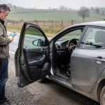 Que faire si une voiture d’occasion tombe en panne après l’achat ? Une voiture d’occasion immobilisée sur le bas-côté d’une route, avec le voyant moteur allumé, tandis qu’un homme consulte le diagnostic de son téléphone près de la portière ouverte.