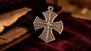 Pendentif en argent orné d’une croix pattée, posé sur un velours sombre à côté d’un parchemin ancien dans une ambiance historique.