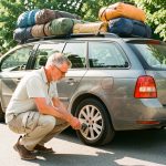 Les 8 vérifications avant un long trajet en voiture Une voiture familiale chargée de bagages stationne sur une aire de repos estivale tandis qu’un homme vérifie ses pneus avant le départ.